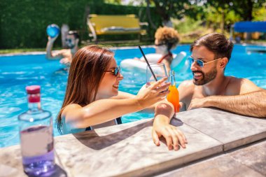 Gençler havuzda güneşli bir yaz gününün tadını çıkarıyorlar. Bir kız ve erkek arkadaşının yüzen ve rahatlatan arkadaşlarıyla birlikte mor kokteyller içip gülmelerine odaklanıyorlar..