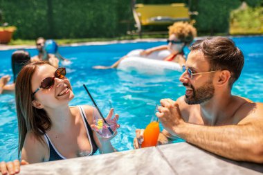 Gençler havuzda güneşli bir yaz gününün tadını çıkarıyorlar. Bir kız ve erkek arkadaşının yüzen ve rahatlatan arkadaşlarıyla birlikte mor ve turuncu kokteyller içip gülmelerine odaklanıyorlar..