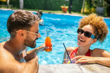 Gençler havuzda güneşli bir yaz gününün tadını çıkarıyorlar; erkek arkadaşıyla yüzen arkadaşlarıyla birlikte mor kokteyl içen afro saçlı kıza odaklanıyorlar..