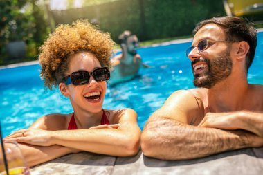 Gençler havuzda güneşli bir günün tadını çıkarıyorlar; afro saçlı kız ve erkek arkadaşının gülümsemesine ve kollarına yaslanmasına odaklanıyorlar, arkadaşlarının yüzdüğü ve arka planda dinlendiği.