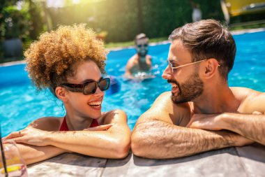 Gençler havuzda güneşli bir günün tadını çıkarıyorlar; afro saçlı kız ve erkek arkadaşının gülümsemesine ve kollarına yaslanmasına odaklanıyorlar, arkadaşlarının yüzdüğü ve arka planda dinlendiği.