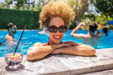 Gençler havuzda güneşli bir yaz gününün tadını çıkarıyorlar; gülümseyen afro saçlı, mor alkollü kokteyl içen, arkadaşlarının yüzdüğü ve arka planda dinlendiği bir kıza odaklanıyorlar..