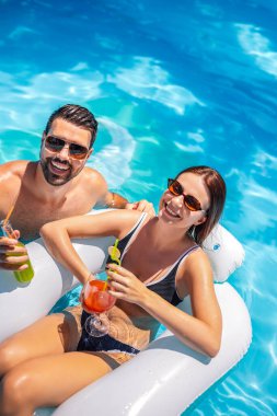 Genç çift güneşli bir yaz gününde havuzda dinleniyor; kız şamandıranın üzerinde, oğlan onun yanında ve ikisi de renkli kokteyller içiyor ve eğleniyorlar..