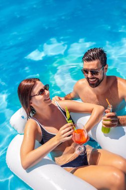 Genç çift güneşli bir yaz gününde havuzda dinleniyor; kız şamandıranın üzerinde, oğlan onun yanında ve ikisi de renkli kokteyller içiyor ve eğleniyorlar..