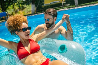 Genç çift havuzda güneşli bir yaz gününün tadını çıkarıyor; afro saçlı kız, partneri yanında renkli kokteyller yudumlarken ve eğlenirken bir şamandıranın üzerinde dinleniyor..