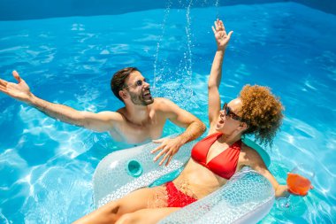 Genç çift havuzda güneşli bir yaz gününün tadını çıkarıyor; afro saçlı kız, partneri yanında renkli kokteyller yudumlarken ve eğlenirken bir şamandıranın üzerinde dinleniyor..