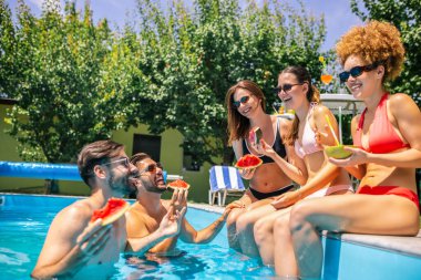 Bir grup genç havuz kenarında takılıyor, bazıları suda bacakları olan, renkli kokteyllerin keyfini çıkaran ve güneşli bir günde birlikte dinlenen insanlar..