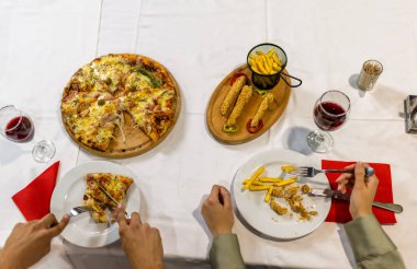 Bir dilim pizza Capricciosa alırken, bir diğeri de yanında bir bardak kırmızı şarapla kızarmış tavuk kesiyor, sıcak bir restoran ortamında, nefis bir yemek anını yakalıyor..