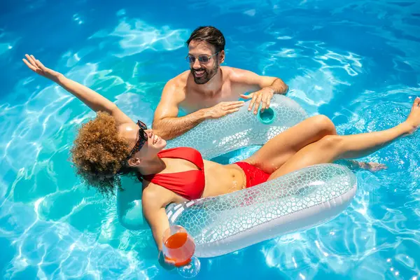 Genç çift havuzda güneşli bir yaz gününün tadını çıkarıyor; afro saçlı kız, partneri yanında renkli kokteyller yudumlarken ve eğlenirken bir şamandıranın üzerinde dinleniyor..