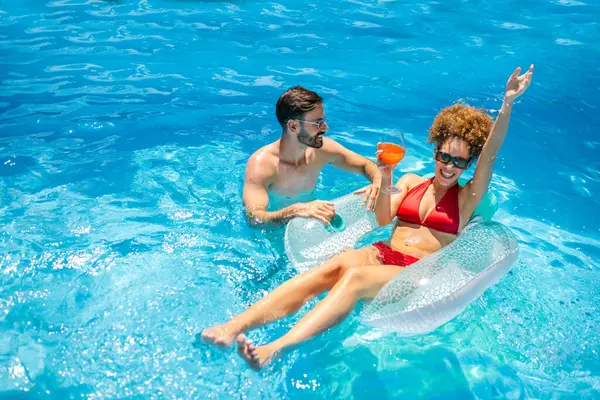Genç çift havuzda güneşli bir yaz gününün tadını çıkarıyor; afro saçlı kız, partneri yanında renkli kokteyller yudumlarken ve eğlenirken bir şamandıranın üzerinde dinleniyor..