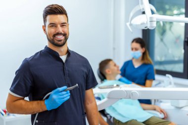 Mavi ceketli erkek dişçi diş aletini tutuyor. Hasta ve başka bir dişçi arka planda bulanıklaşıyor. Bakım, tedavi ve profesyonel diş hizmetleri kavramı.