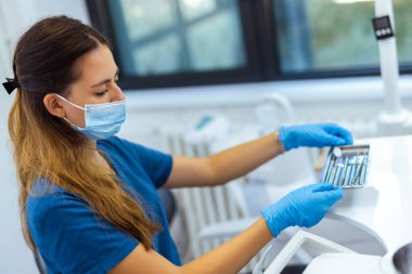 Modern klinikte mavi ceket ve yüz maskesi takan kadın dişçi diş aletlerini hazırlıyor. Hijyen, hazırlık ve profesyonel bakım kavramı