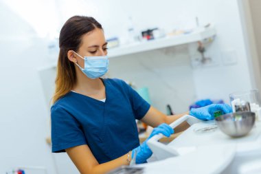 Modern klinikte mavi ceket ve yüz maskesi takan kadın dişçi diş aletlerini hazırlıyor. Hijyen, hazırlık ve profesyonel bakım kavramı