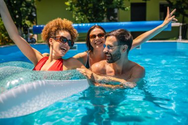 Genç bir kadın havuzda şişme bir yüzüğün üzerinde yüzerken gülerken arkadaşları onu şakacı bir şekilde itmiş. Arkadaşlarla eğlenceli bir yaz anıydı. Suyun ve güneşin tadını çıkarıyordu..