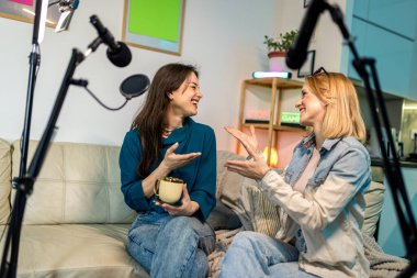 Evde podcast yapan iki genç kadın, kahve içip sohbet ediyorlar. Etrafı mikrofonlar, ışıklar ve kameralarla çevrili..