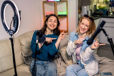 Evde podcast yapan iki genç kadın ellerini hareket ettirirken gülüyor ve dans öğreniyorlar. Mikrofonlar, ışıklar ve çevrelerinde kamera var..