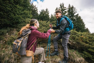 Tam teçhizatlı bir yürüyüş çifti, bir dağ yoluna tırmanır, erkek, nefes kesici manzarayla çevrili, kadının elini tutarak kadına yardım eder..