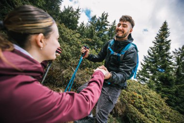 Tam teçhizatlı bir yürüyüş çifti, bir dağ yoluna tırmanır, erkek, nefes kesici manzarayla çevrili, kadının elini tutarak kadına yardım eder..
