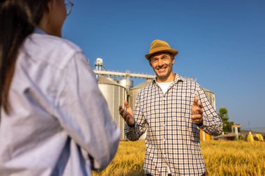 İki işçi buğday kaplı bir alanda büyük siloların önünde duruyor ve gelecek görevleri aktif olarak tartışıyorlar..