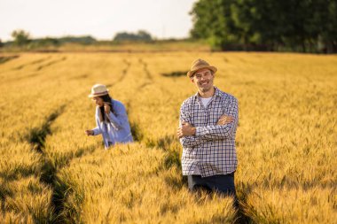 Üst düzey bir adam altın buğday tarlasında dikilirken kameraya güvenle gülümsüyor. Arka planda özenle çalışan Taylandlı bir kadın çiftçi ile birlikte..