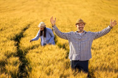 Üst düzey bir adam altın buğday tarlasında dikilirken kameraya güvenle gülümsüyor. Arka planda özenle çalışan Taylandlı bir kadın çiftçi ile birlikte..