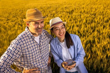 Taylandlı bir kadın ve yaşlı bir erkek işçi büyük siloların önünde bir buğday yığınının üzerinde, akıllı telefon kullanarak birlikte duruyorlar..