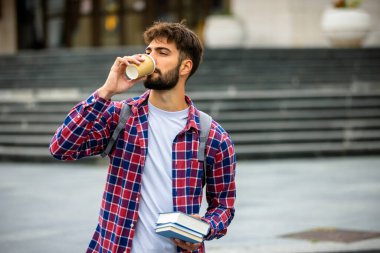 Ekose gömlekli genç erkek öğrenci kitap okuyor ve üniversite kampüsünde dışarıda otururken kahve içmekten zevk alıyor..