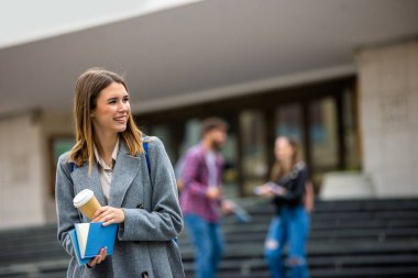 Üniversite kampüsünün dışında elinde kitap ve kahveyle duran genç bir kız öğrenci ve arka planda bulanık meslektaşları var..