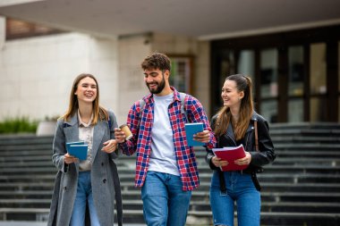 Üç öğrenci, bir erkek ve iki kadın. Üniversite kampüsünün dışında birlikte gülüyorlar. Ders arasında ellerinde kitaplarla..