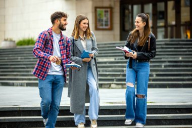 Üç öğrenci, bir erkek ve iki kadın. Üniversite kampüsünün dışında birlikte gülüyorlar. Ders arasında ellerinde kitaplarla..