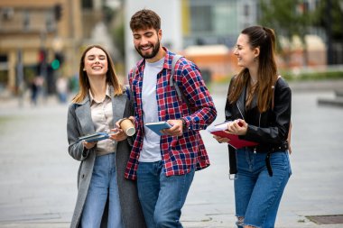 Üç öğrenci, bir erkek ve iki kadın. Üniversite kampüsünün dışında birlikte gülüyorlar. Ders arasında ellerinde kitaplarla..