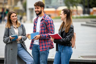 Üç öğrenci, bir erkek ve iki kadın. Üniversite kampüsünün dışında birlikte gülüyorlar. Ders arasında ellerinde kitaplarla..