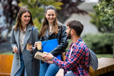 Üç öğrenci, bir erkek ve iki kadın, kampüsün dışında ellerinde kitaplarla gülüyorlar. Bu arada erkek, ders arası için kahve getiriyor..