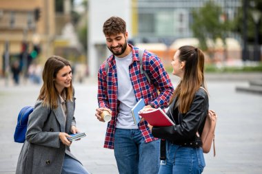 Üç öğrenci, bir erkek ve iki kadın. Üniversite kampüsünün dışında birlikte gülüyorlar. Ders arasında ellerinde kitaplarla..