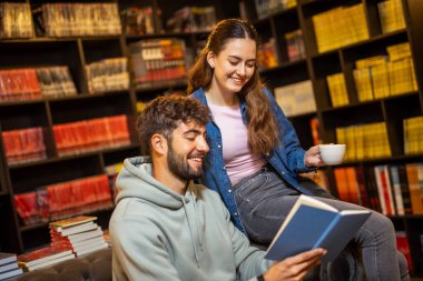 Genç bayan ve erkek öğrenciler rahat bir kütüphane kafesinde, kitaplarla çevrili bir şekilde kahve içerken birlikte kitap okuyorlar..