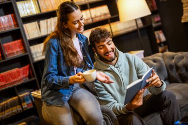 Genç bayan ve erkek öğrenciler rahat bir kütüphane kafesinde, kitaplarla çevrili bir şekilde kahve içerken birlikte kitap okuyorlar..