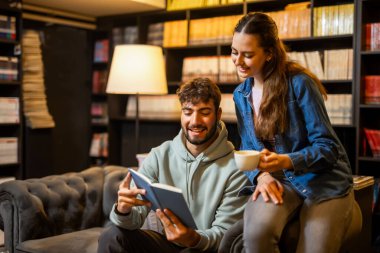 Genç bayan ve erkek öğrenciler rahat bir kütüphane kafesinde, kitaplarla çevrili bir şekilde kahve içerken birlikte kitap okuyorlar..