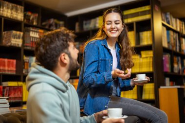 Genç bayan ve erkek öğrenciler kütüphanedeki kafede oturuyor, kız kanepede oturuyor, oğlan kanepede oturuyor, kahve içiyor ve birlikte sohbet ediyorlar..