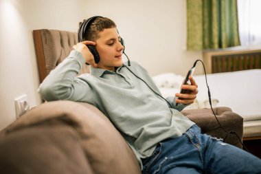 Yolculukta olan genç bir çocuk, bir dairede tek başına, kulaklık takıyor ve akıllı telefonuyla müziğin keyfini çıkarıyor, evde dinleniyor..