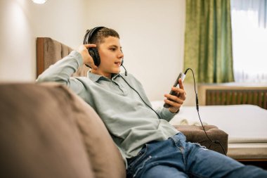 Yolculukta olan genç bir çocuk, bir dairede tek başına, kulaklık takıyor ve akıllı telefonuyla müziğin keyfini çıkarıyor, evde dinleniyor..