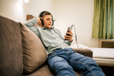 Yolculukta olan genç bir çocuk, bir dairede tek başına oturuyor, kulaklık takıyor ve akıllı telefonunu kullanıyor, müzik ve oyunlardan hoşlanıyor..