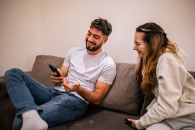 Genç bir çift otel odasında tatilde, erkek arkadaşı içeride dinlenirken kız arkadaşına telefonunda bir şey gösteriyor..