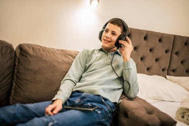 Yolculukta olan genç bir çocuk, bir dairede tek başına, kulaklık takıyor ve akıllı telefonuyla müziğin keyfini çıkarıyor, evde dinleniyor..