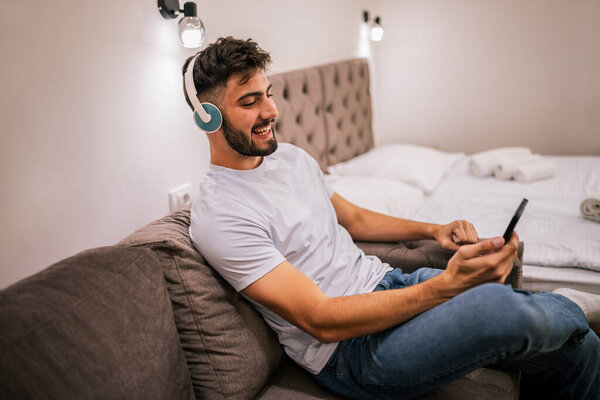 Мужчина сидит один в гостиничном номере, в наушниках, смотрит что-то на смартфоне и наслаждается развлечениями в помещении.