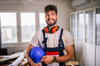 Kask ve kulaklık takan tamirci, elinde bir inşaat planı tutarken kameraya gülümsüyor. .