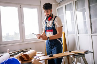 Tamirci apartman zeminini onarıyor, etrafı lamine tahtalarla çevrili, ve rehberlik için bir tablet kullanıyor, modern teknolojiyi ustalıkla birleştiriyor..