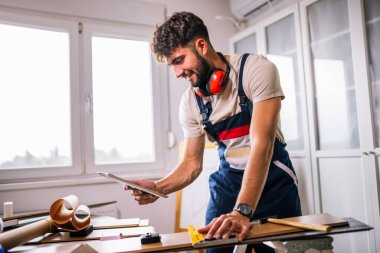 Tamirci apartman zeminini onarıyor, etrafı lamine tahtalarla çevrili, ve rehberlik için bir tablet kullanıyor, modern teknolojiyi ustalıkla birleştiriyor..