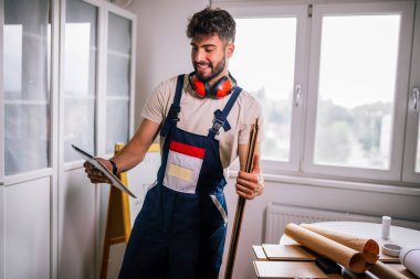 Tamirci apartman zeminini onarıyor, etrafı lamine tahtalarla çevrili, ve rehberlik için bir tablet kullanıyor, modern teknolojiyi ustalıkla birleştiriyor..