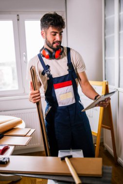 Tamirci apartman zeminini onarıyor, etrafı lamine tahtalarla çevrili, ve rehberlik için bir tablet kullanıyor, modern teknolojiyi ustalıkla birleştiriyor..