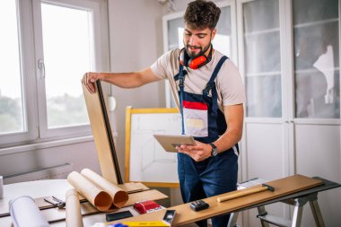 Tamirci apartman zeminini onarıyor, etrafı lamine tahtalarla çevrili, ve rehberlik için bir tablet kullanıyor, modern teknolojiyi ustalıkla birleştiriyor..